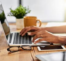 Hands typing on a laptop at a wooden desk with glasses, a notebook, a smartphone, and a coffee mug nearby.
