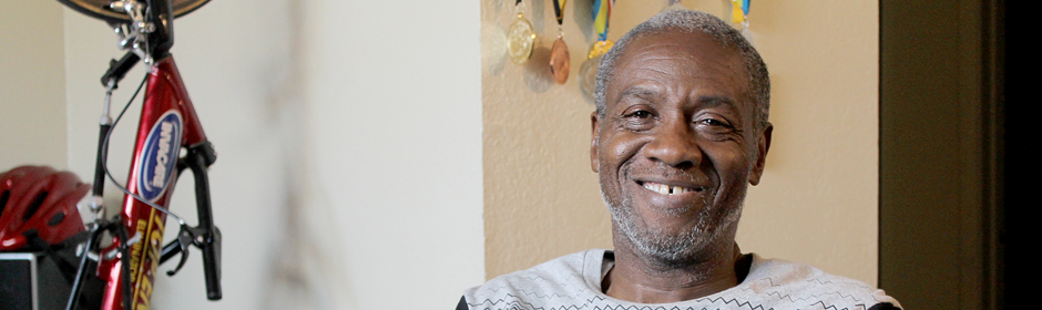 Smiling older man seated indoors with medals on the wall and a bicycle frame nearby.