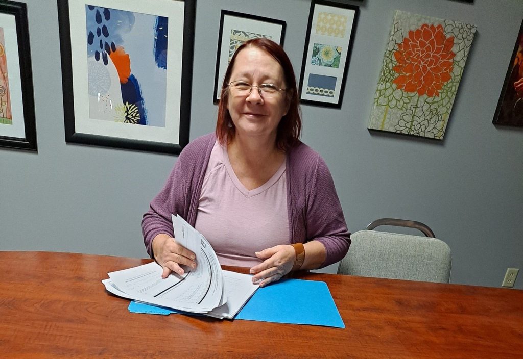 Woman in pink top and purple cardigan sits at a wooden table, flipping through papers in an art-filled room.