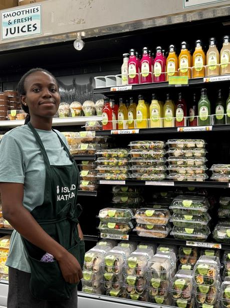 Grocery store worker in a green apron stands beside a refrigerated display of colorful juices and pre-packaged salads.