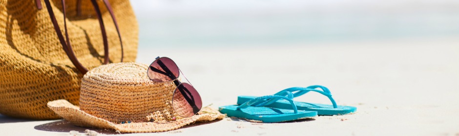 A straw sunhat with sunglasses resting on the sand beside turquoise flip-flops at the beach.