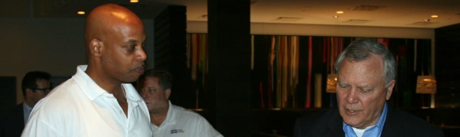 Two men converse in a dimly lit room, one bald in a white polo and the other with gray hair in a dark blazer.