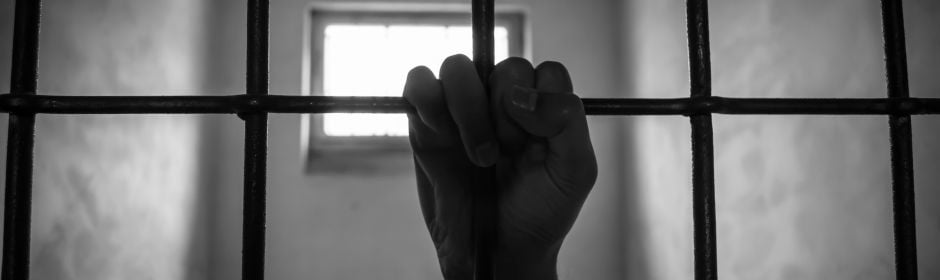 A hand tightly grips metal prison bars inside a dim, empty cell.