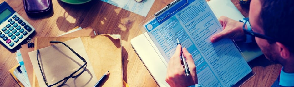 Person fills out a form on a clipboard at a desk, with glasses, a calculator, and scattered papers.