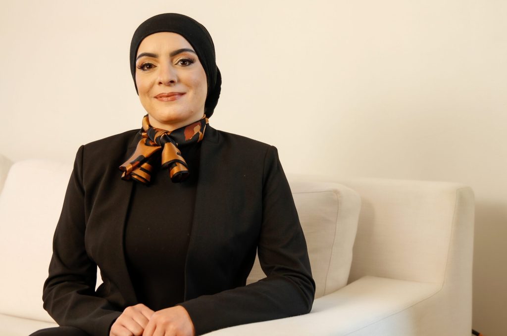 Professional woman in a black hijab and blazer seated on a white sofa.