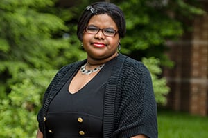 Black woman wearing a black cardigan and pearl necklace stands outdoors among green shrubs.