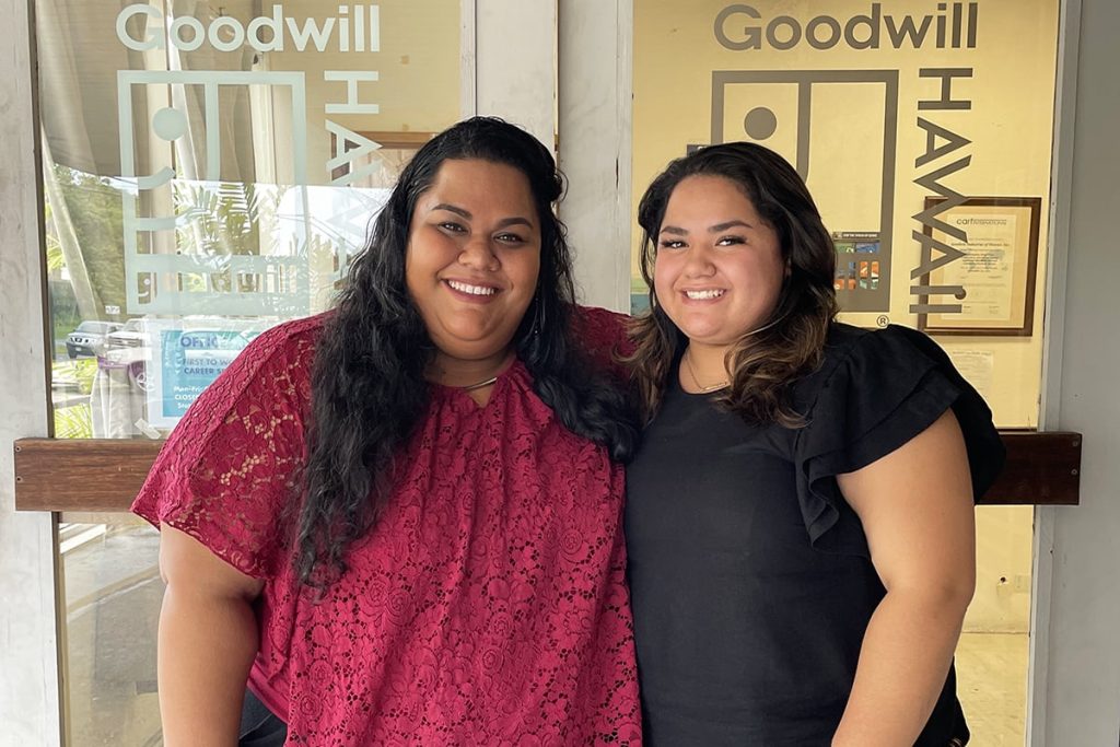 Two women smile and pose together in front of a Goodwill storefront window.