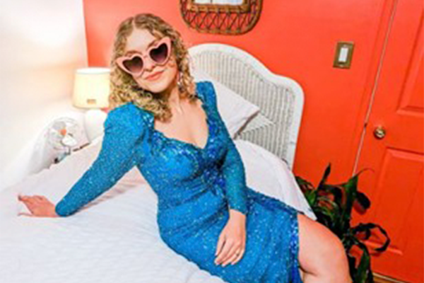 Woman with curly blonde hair wearing a blue sequin dress and pink heart-shaped sunglasses lounging on a bed.