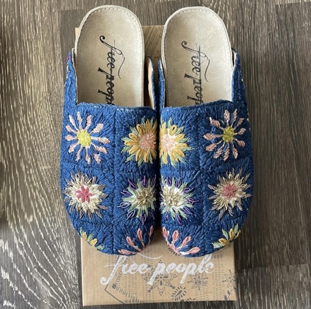 Blue velvet slip-on mules featuring multicolored daisy embroidery, displayed on a wooden floor with a Free People box.