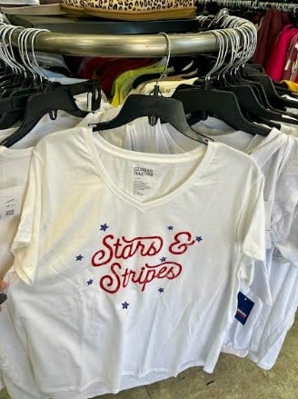 White T-shirt on display featuring a red 'Stars & Stripes' cursive logo with blue stars on a clothing rack.
