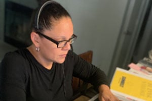 Woman wearing glasses sits at a desk, focused on documents with a yellow booklet nearby.