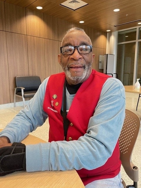 Smiling older man with gray beard and glasses, wearing a red vest over a light blue shirt, seated at a table in a wood-paneled room.