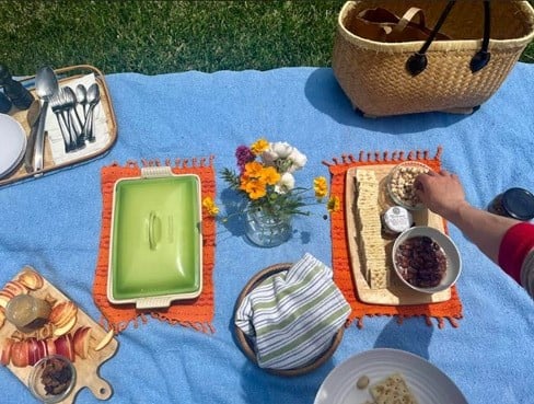 An image of a pinic setup in a garden with a purse, a cheese and crackers tray and flowers on top of a blue blanket