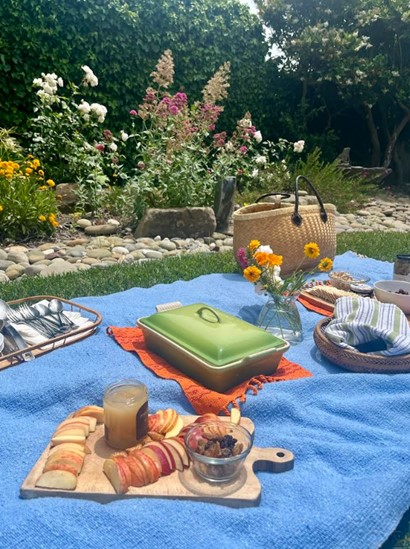 Picnic setup on a blue blanket in a garden with sliced bread, jam, a green lunchbox, flowers, and a wicker basket.
