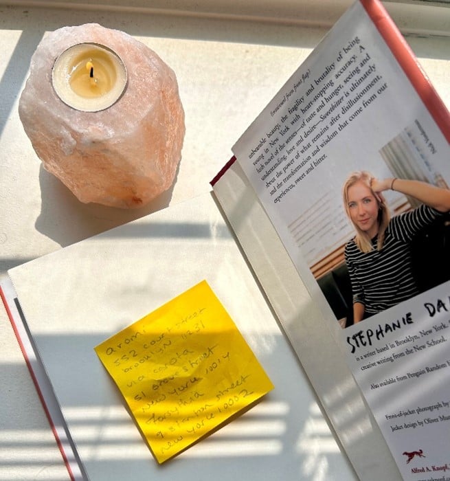 Pink Himalayan salt lamp with a lit candle beside an open magazine and a yellow sticky note on a sunlit white surface.