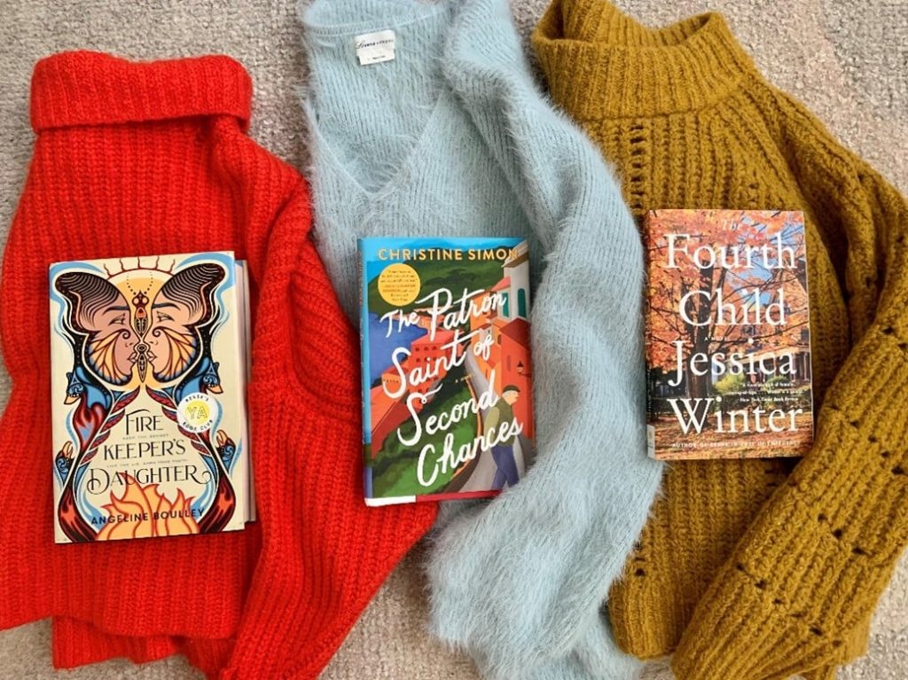Three knit sweaters in red, light blue, and mustard with three novels resting on top.
