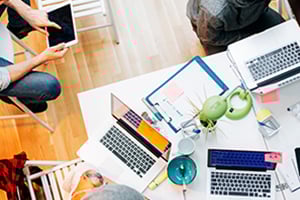 Overhead view of a collaborative workspace with laptops, tablets, papers, and colorful stationery around a white table.