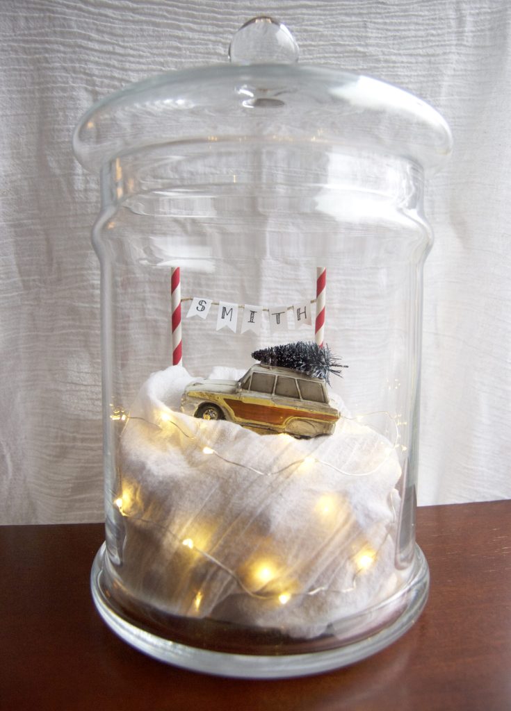 Miniature car with a tiny Christmas tree on its roof sits on a snowy base inside a glass cloche, surrounded by candy-cane poles and fairy lights.