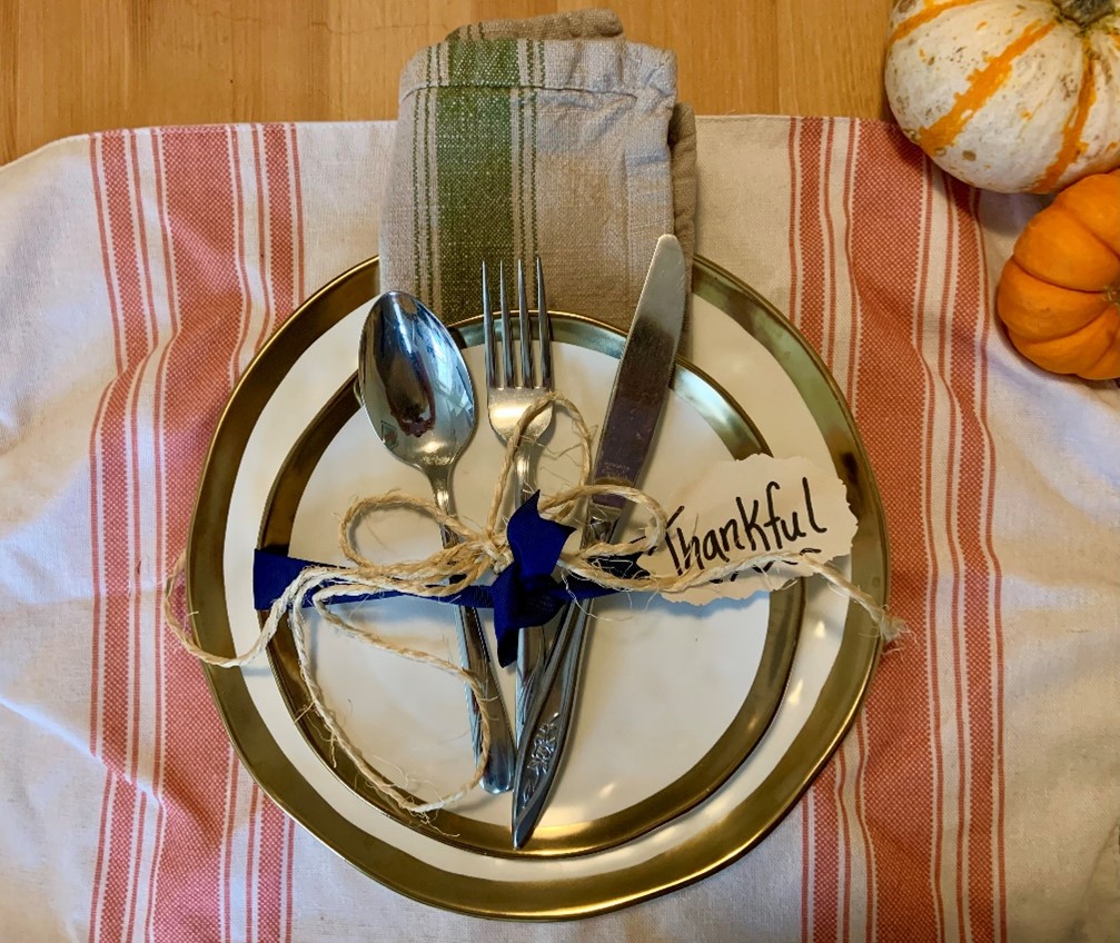 Table setting with gold-rimmed plates, tied cutlery, and a 'thankful' tag on a striped tablecloth, with pumpkins nearby.