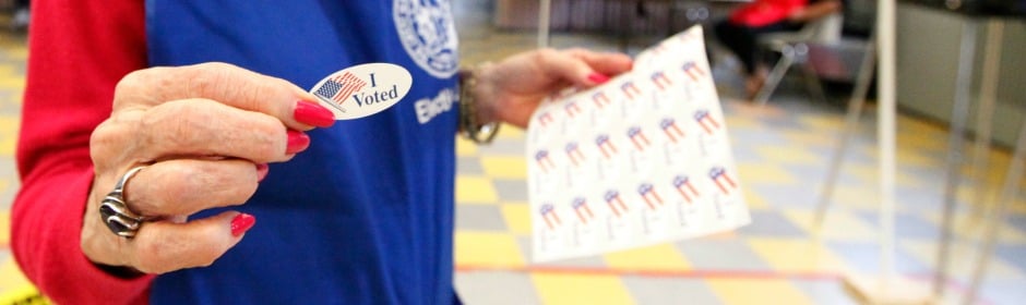 Close-up of a hand with red nails and a ring, holding an 'I Voted' sticker and a sheet of stickers in a public indoor space.