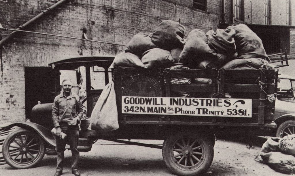 Black-and-white image of a vintage Goodwill truck loaded with burlap sacks, with a man standing beside the cab.