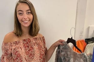 Smiling young woman with shoulder-length hair wearing an off-shoulder floral top beside a clothing rack.