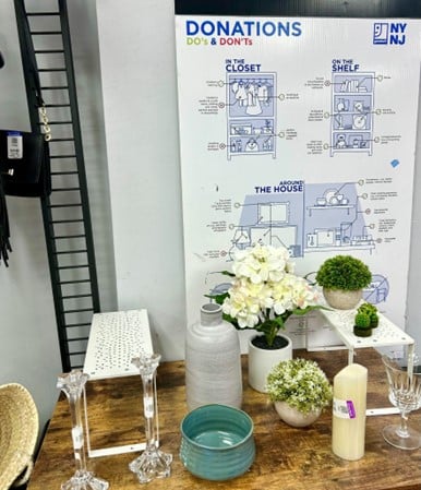 Donation station display with a 'DONATIONS DO's & DON'Ts' poster on the wall and a wooden table showcasing white hydrangea flowers, small plants, glassware, and a candle.