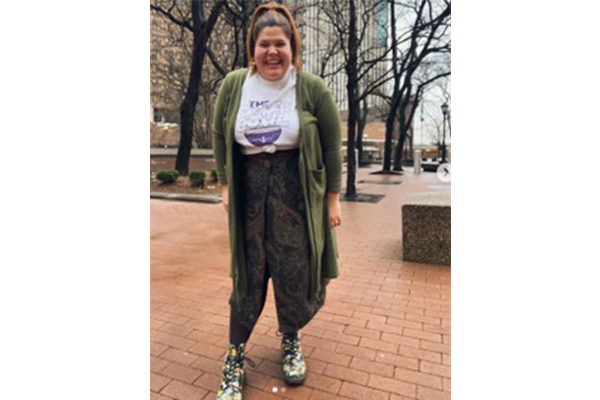 Smiling woman in a green cardigan, white T-shirt, and paisley pants posing outdoors on a brick walkway.