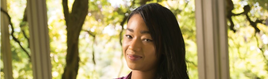 Portrait of a smiling woman with long dark hair outdoors, with sunlit trees in the background.