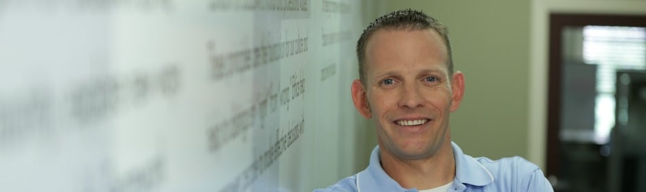 Smiling man in a light blue polo stands beside a glass wall with blurred text in an office.