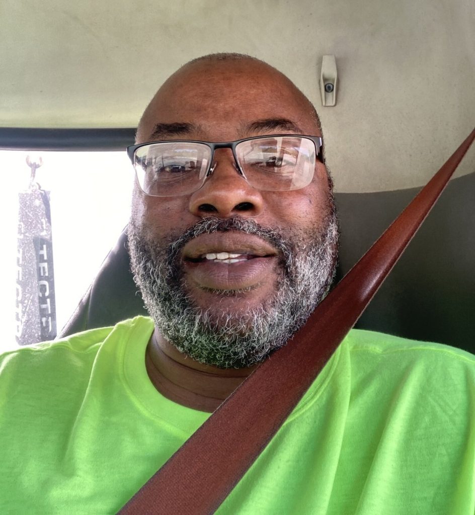 Smiling man with a gray beard and glasses wearing a bright lime-green shirt, secured with a seat belt in a car.