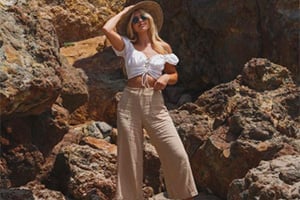 Woman in white crop top and beige wide-leg pants posing among rocky terrain under sunny sky, wearing hat.