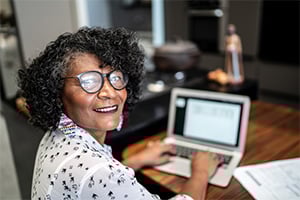 Smiling woman with curly hair and glasses using a laptop at a desk in an office.