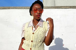 Woman in pale yellow knit vest with a pink lace collar and orange sunglasses, standing against a white wall with a strong shadow.