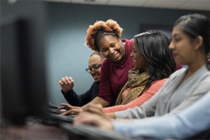 Group of diverse students working on laptops in a computer lab, smiling as they collaborate