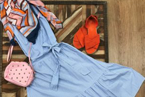 Blue striped belted dress laid on a wooden floor, beside a pink perforated purse and orange sandals.