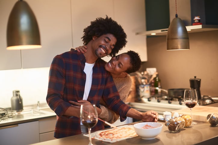Couple smiling and embracing in a modern kitchen as they prepare pizza.