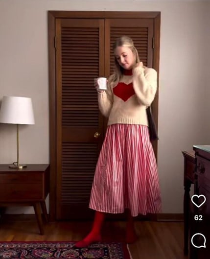 Young woman in a cream sweater with a red heart, red tights, and striped skirt, holding a mug indoors.