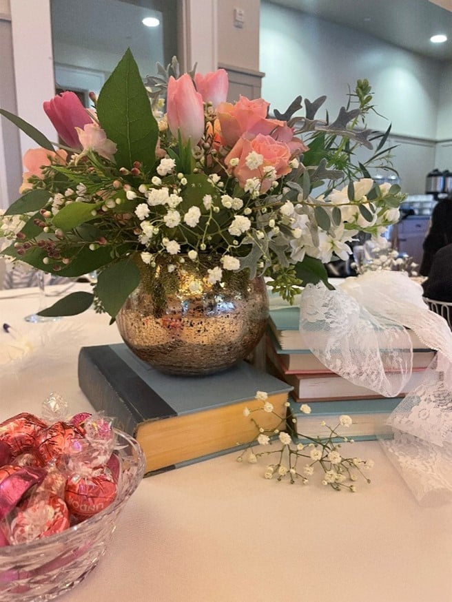 Bouquet of pink tulips and white baby's breath in a metallic vase atop a stack of books on a banquet table.