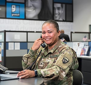 Female soldier in camouflage uniform sits at a desk, talking on a phone in a busy office.