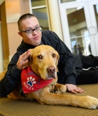 Man with glasses pets a tan service dog wearing a red paw-print bandana.