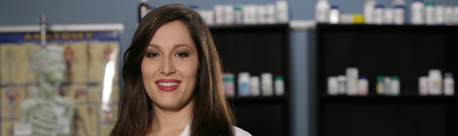 Female pharmacist in a lab coat smiles in a pharmacy with shelves of medication behind.