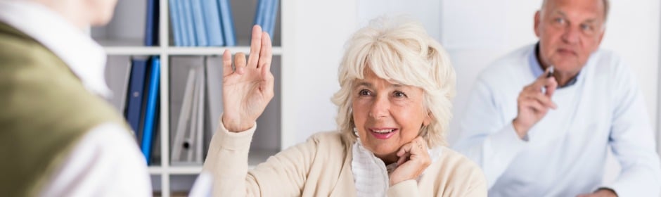 Elderly woman raises her hand in a discussion with colleagues in an office setting.