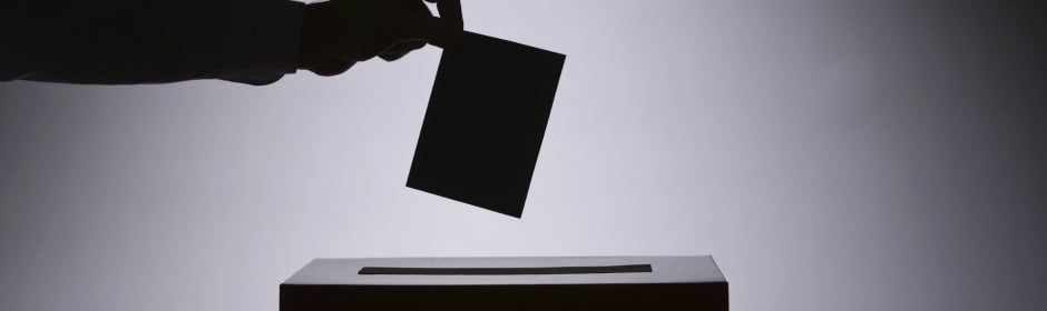Silhouette of a hand dropping a ballot into a ballot box.
