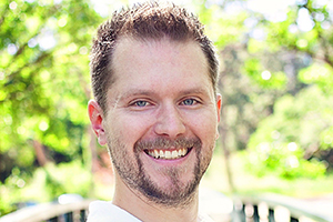 Smiling man with short brown hair and stubble outdoors in bright daylight.