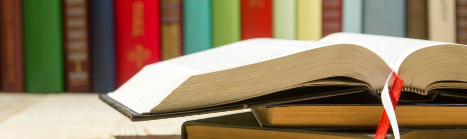 Open book with a red bookmark resting on a stack of books, colorful spines visible in the background.