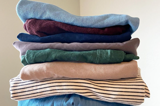 Stack of folded clothes in pastel colors (light blue, burgundy, navy, gray, beige, striped) arranged on a surface.