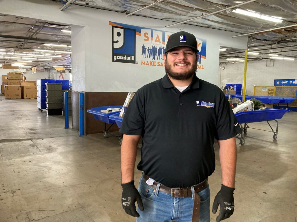 Smiling Goodwill employee in a black polo and cap stands in a warehouse with blue sorting bins and pallets.