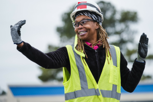 Construction worker wearing a neon hi-visibility vest and helmet, smiling with arms raised at an outdoor site.