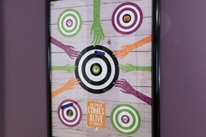 Framed poster with a large central bullseye surrounded by colorful circular targets and several arms reaching toward the center.